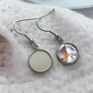 May include: A pair of silver earrings with a round, clear glass charm. One charm has a pink and gold abstract design, while the other charm is blank.