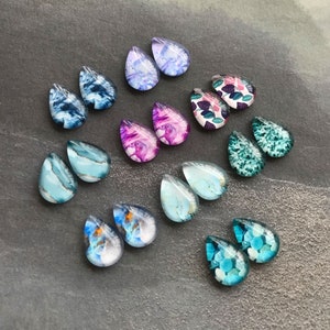 Op de afbeelding: Een verzameling druppelvormige oorbellen in verschillende kleuren en patronen. De oorbellen hebben ontwerpen in tinten blauw, paars, roze en groen, met gemarmerde, bloemen- en abstracte patronen. De oorbellen worden getoond op een donkergrijze ondergrond.