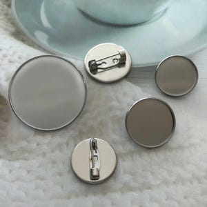 May include: A selection of blank, circular, silver-coloured pinback buttons in different sizes. Two buttons have pin backs attached, while the others are plain. The buttons are on a white, textured surface, with a light blue teacup in the background.