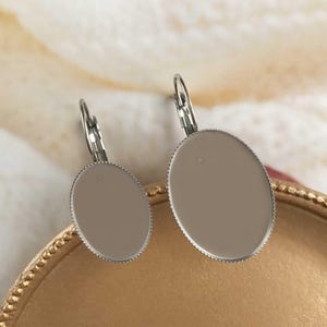 Könnte beinhalten: Ein Paar ovale Ohrringe mit hellbraunem Mittelteil und silberfarbenem, strukturiertem Rand. Die Ohrringe haben einen Klappbügelverschluss. Die Ohrringe werden auf einer goldfarbenen Oberfläche präsentiert.