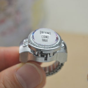 May include: Silver-toned ring with a domed, circular face. The face features the words "DREAMS COME TRUE" in black text against a white background, surrounded by a floral design. The ring has a textured bezel and an adjustable band.