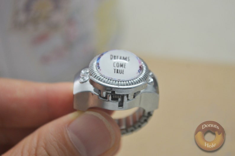 May include: Silver-toned ring with a domed, circular face. The face features the words "DREAMS COME TRUE" in black text against a white background, surrounded by a floral design. The ring has a textured bezel and an adjustable band.