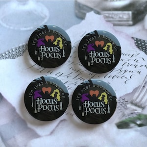 Puede incluir: Cuatro botones redondos negros con fondo blanco y el texto "It's just a bunch of Hocus Pocus" con tres siluetas de brujas en el centro. Las brujas son moradas, naranjas y amarillas.