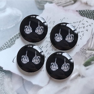 May include: Four black round buttons each featuring a white skeleton hand giving the middle finger.