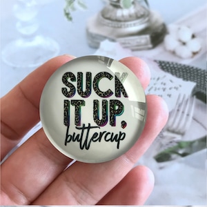 May include: A round glass button with a white background and black text that reads "SUCK IT UP, buttercup". The text is in a leopard print pattern with rainbow iridescent accents.