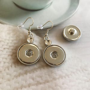 May include: A pair of silver earrings with clear rhinestone accents and a silver button charm. The earrings are on a white surface.
