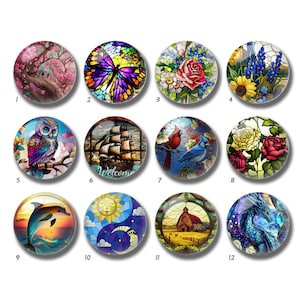 May include: A collection of twelve round glass cabochons with colorful stained-glass designs. The designs include a cherry blossom tree, a butterfly, roses, sunflowers, an owl, a sailboat, cardinals, a dolphin, a sun and moon, a barn, and a dragon.