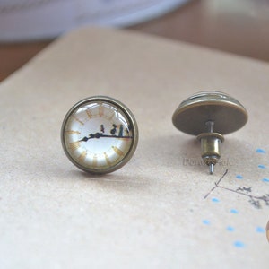 May include: A pair of bronze stud earrings with a round glass cabochon featuring a vintage clock face with Roman numerals and a silhouette of people walking across the face.