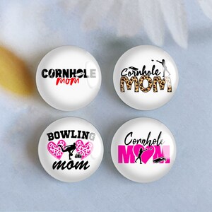 May include: Four white round buttons with different designs. The buttons feature the text "Cornhole Mom" with different graphics. One button has a black and white graphic of a person throwing a bean bag. Another button has a leopard print background. The third button has a pink heart with a black and white graphic of a person bowling. The fourth button has a pink heart with a black and white graphic of a person throwing a bean bag.