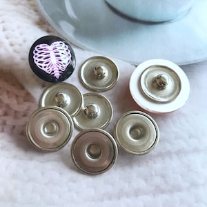 May include: A set of seven silver-toned snap buttons, with one button featuring a pink and black heart-shaped design with a ribcage graphic.