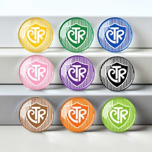 May include: A set of nine circular glass cabochons, each featuring a white shield emblem with the letters CTR in the center. The background of each shield consists of vertical stripes in yellow, green, blue, pink, purple, black, brown, orange, and lime.