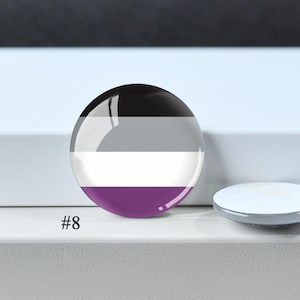 Puede incluir: Un bot&oacute;n circular con alfiler que muestra la bandera del orgullo asexual con franjas horizontales en negro, gris, blanco y morado. El bot&oacute;n tiene un acabado brillante y est&aacute; etiquetado con el n&uacute;mero 8 en la esquina inferior izquierda.