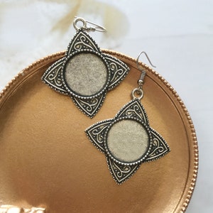 Puede incluir: Un par de pendientes de plata con un diseño en forma de estrella y un espacio redondo en blanco en el centro. Los pendientes están sobre una superficie de color dorado.