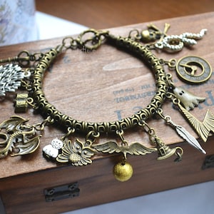Puede incluir: Un brazalete de charms de bronce con una variedad de charms, incluyendo un dragón, un búho, una pluma, un rayo, un fantasma, un reloj, un árbol, un sombrero y una campana. El brazalete está sobre una superficie de madera.