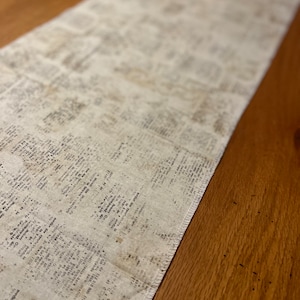 Könnte beinhalten: Beiger Tischläufer mit Zeitungsmuster. Der Stoff ist cremefarben mit verblasstem schwarzem Text, was ihm einen Vintage-Look verleiht. Der Läufer liegt auf einer Holzoberfläche.