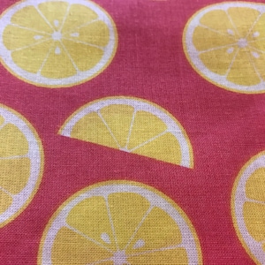 May include: A close-up of pink fabric featuring a repeating pattern of yellow lemon slices. Each lemon slice is outlined in white, creating a bright design. The fabric has a textured appearance.