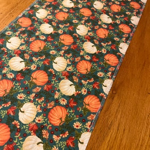 May include: A teal table runner with a repeating pattern of orange and white pumpkins, fall leaves, and small white flowers. The runner has a stitched edge and is displayed on a wooden table.