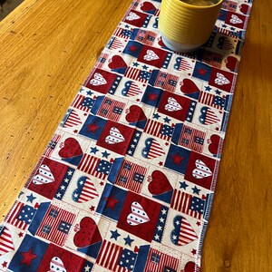 May include: A patriotic table runner with a repeating pattern of hearts, stars, and American flags in red, white, and blue. A yellow mug with a brown liquid sits on the runner, which is placed on a wooden table.
