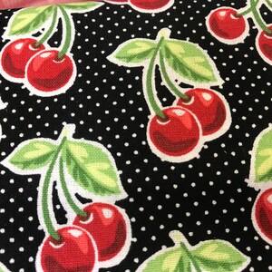 May include: Close-up of a fabric featuring a repeating pattern of red cherries and green leaves on a black background with white polka dots. The cherries have a white outline.