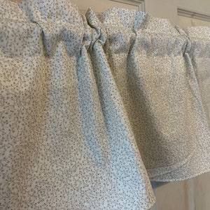 May include: A set of white valance curtains with a delicate gray floral pattern. The fabric is gathered at the top, creating soft folds. The curtains are designed to be hung on a rod above a window.