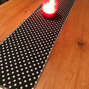 Puede incluir: Un camino de mesa de lunares blancos y negros con una vela roja en un portavelas de cristal. El camino de mesa es rectangular y está colocado sobre una mesa de madera. La vela está encendida, emitiendo un brillo cálido.