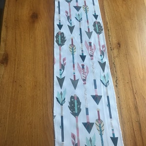 May include: A white fabric runner with a repeating pattern of arrows in shades of gray, pink, teal, and yellow. The arrows have decorative feather and leaf details. The runner is rectangular and appears to be made of cotton or a similar fabric.