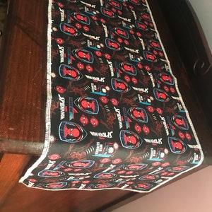 Puede incluir: Camino de mesa negro con un patrón repetido de logotipos de Spider-Man, arte de cómic y texto. El diseño incluye acentos rojos, azules y blancos. El camino de mesa tiene un borde blanco con pespunte.