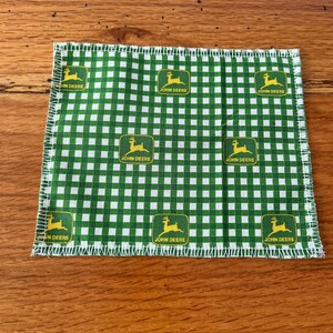 May include: A green and white checkered cloth with a repeating pattern of the John Deere logo. The logo features a yellow deer inside a green rectangle. The cloth has a white stitched edge.