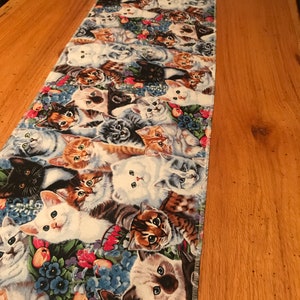 May include: A rectangular table runner featuring a repeating pattern of various cat breeds. The cats are depicted in a variety of colors, including white, black, brown, and gray, with floral accents in blue, pink, and green. The runner is made of fabric.