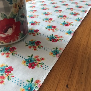 May include: White floral table runner with a repeating pattern of red, blue, and yellow flowers. The runner has a dotted border and a decorative edge. A floral-patterned ceramic jar is partially visible on the left.