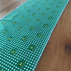 May include: Green and white checkered table runner with a repeating pattern of the John Deere logo. The runner is made of fabric and has a raw edge. The runner is placed on a wooden table.