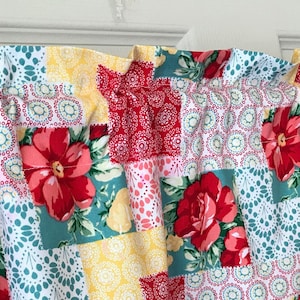 Op de afbeelding: Een patchwork valance met een bloemenpatroon. De valance heeft een herhalend patroon van rode, gele en blauwgroene vierkanten met grote rode bloemen. De stof is aan de bovenkant gerimpeld, waardoor een gerimpeld effect ontstaat. De valance is waarschijnlijk voor een keuken- of slaapkamerraam.