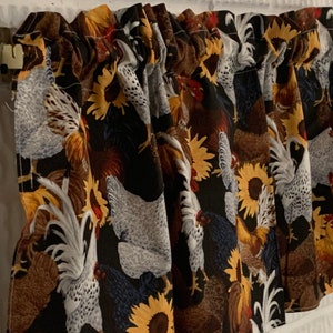 May include: A valance curtain with a black background featuring a repeating pattern of chickens and sunflowers in shades of brown, yellow, and white. The curtain is mounted on a white brick wall.