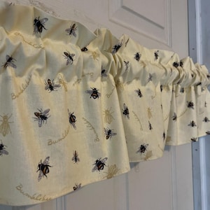 Op de afbeelding: Crèmekleurige valance met een herhalend patroon van zwarte en gele hommels en het woord "Bumblebee" in een scriptlettertype. De stof is aan de bovenkant geplooid, waardoor zachte plooien ontstaan.