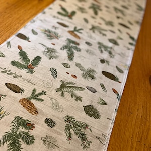 Può includere: Runner da tavolo rettangolare beige con un motivo ripetuto di rami di pino verdi, pigne e illustrazioni botaniche. Il runner ha un bordo cucito bianco ed è appoggiato su una superficie di legno.