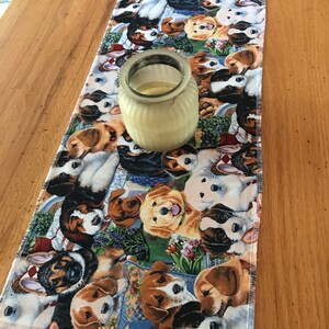 May include: A rectangular table runner featuring a colorful pattern of various dog breeds. A small, clear glass jar with a light-colored substance inside sits on the runner. The runner's design includes dogs of different colors and sizes.