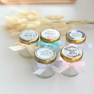 May include: Five small glass jars with gold lids and floral themed labels. The labels have text that says "Thank you for showering" followed by a name, and "with love". One jar has a label that says "Ashwini's Baby Shower".