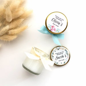May include: Two small glass jars with white candles and gold lids. The lids have floral patterned labels with the text "Thank you for showering Olivia's with love!" and "Thank you for showering Jacob's with love!" The jars are tied with blue and white ribbons.