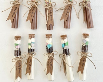May include: A set of ten glass test tubes filled with various ingredients for making hot chocolate. Each tube is tied with twine and a brown paper tag that reads "Baby Alert" and the date "MAY 2023".