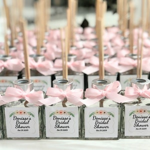 Pode incluir: Uma coleção de pequenas garrafas de vidro com tampas prateadas e laços cor-de-rosa. Cada garrafa contém um líquido transparente e um pau de madeira. As garrafas têm etiquetas que dizem "Denisse's Bridal Shower Oct 19, 2019".