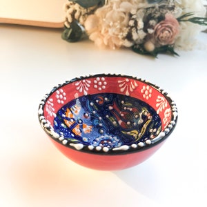 May include: A small, round ceramic bowl with a red exterior and a detailed blue interior. The rim is decorated with white dots and floral patterns. The bowl is likely used for serving food or as a decorative piece.