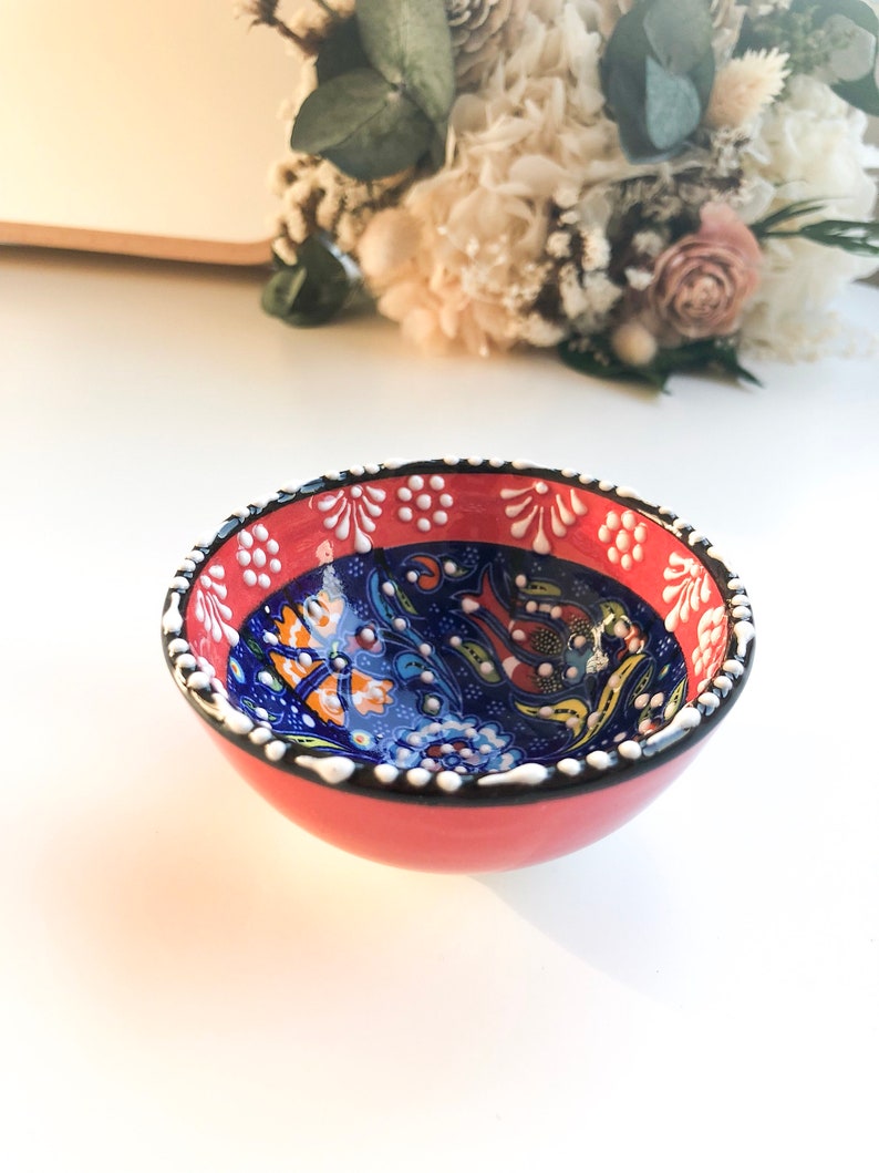 May include: A small, round ceramic bowl with a red exterior and a detailed blue interior. The rim is decorated with white dots and floral patterns. The bowl is likely used for serving food or as a decorative piece.