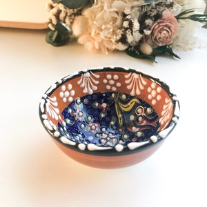 May include: A small, round ceramic bowl with a hand-painted floral design. The bowl features a brown exterior, a blue interior with colorful flowers, and a white and black rim. The bowl is likely used for serving food or as a decorative piece.