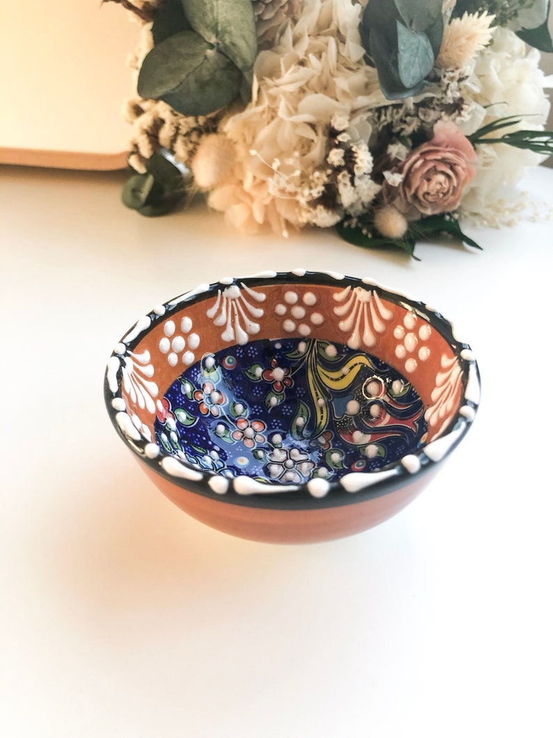 May include: A small, round ceramic bowl with a hand-painted floral design. The bowl features a brown exterior, a blue interior with colorful flowers, and a white and black rim. The bowl is likely used for serving food or as a decorative piece.