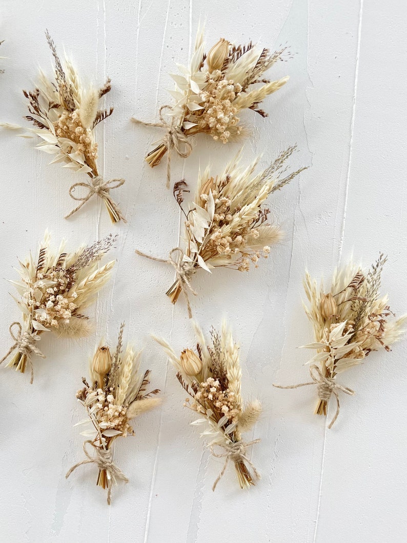 May include: Seven small bouquets of dried flowers in shades of beige and brown, tied with twine. The bouquets are arranged on a white textured background.