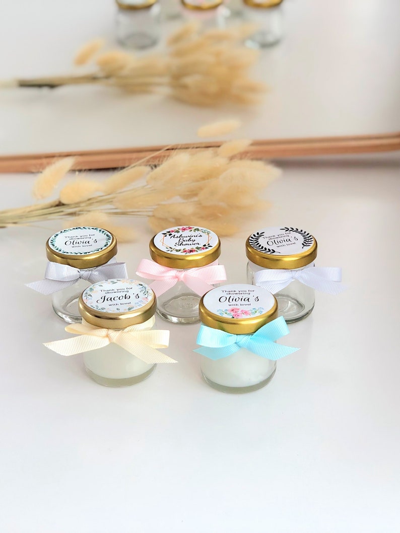 May include: Six small glass jars with gold lids and personalized labels. The labels have floral designs and text that says "Thank you for celebrating with love" and the names "Olivia's", "Jacob's", "Olivia's", and "Ashwini's Baby Shower". The jars are tied with different colored ribbons.