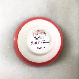 May include: A small, round, ceramic dish with a red rim and white interior. The dish features a sticker with the text "Caitlin's Bridal Shower" and the date "01.19.19" along with floral illustrations. The dish is likely a party favor.