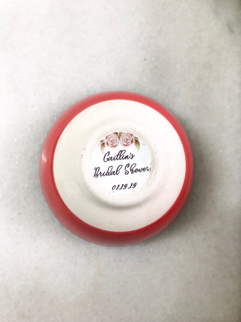 May include: A small, round, ceramic dish with a red rim and white interior. The dish features a sticker with the text "Caitlin's Bridal Shower" and the date "01.19.19" along with floral illustrations. The dish is likely a party favor.
