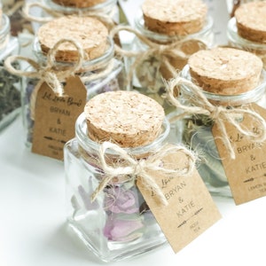 May include: A collection of small glass jars with cork lids, filled with dried flowers and tied with twine. Each jar has a brown paper tag with the text "Let Love Brew Bryan & Katie" and the type of tea.