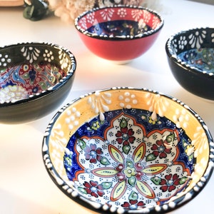 May include: Four small, colorful ceramic bowls with intricate floral patterns. The bowls have a variety of colors, including red, black, yellow, and blue. The interior designs feature detailed floral motifs, and the rims are decorated with white dots.